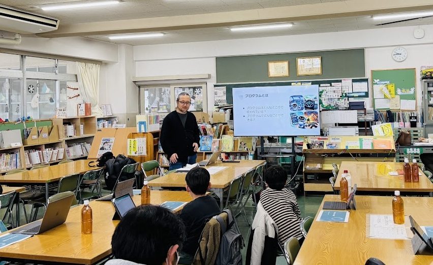 小学校の教室で、スクリーンに「プログラムとは」のスライドを映して説明している体験会の様子